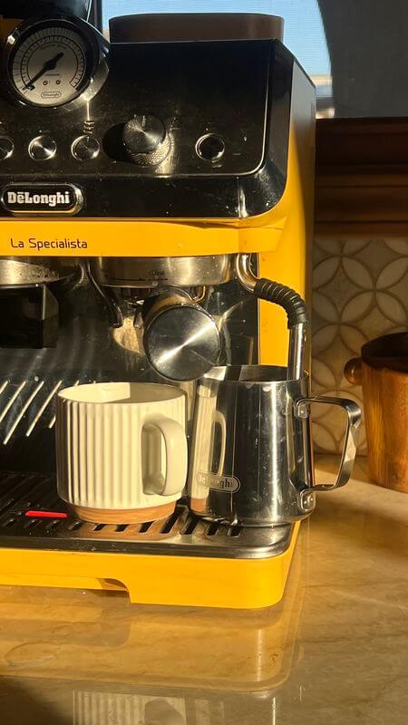 מכונת קפה צהובה קטנה מוארת באור בוקר טבעי, עם כוס לבנה וקנקן נירוסטה